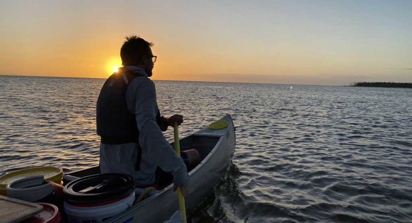 A person at the front of a canoe paddles the boat on calm water. The sun appears to be setting behind their head. 